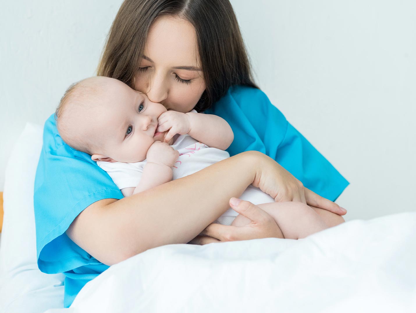 Una mamá vestida con una bata azul, cargando a su bebé recién nacido y besándolo en la mejilla izquierda