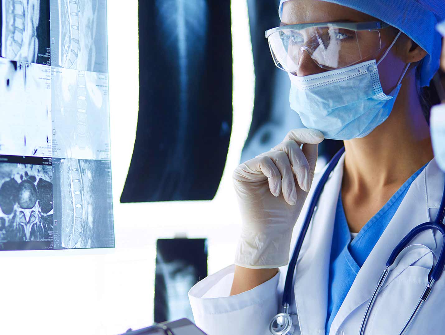 Doctora con bata blanca, cubrebocas, gorro quirúrgico y guantes blancos, observando detenidamente una radiografía