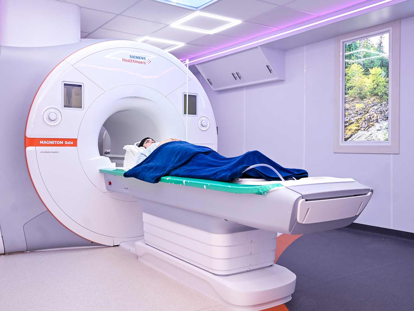Paciente acostada y tapada con una manta azul en la camilla del equipo de resonancia magnética de color blanco con naranja