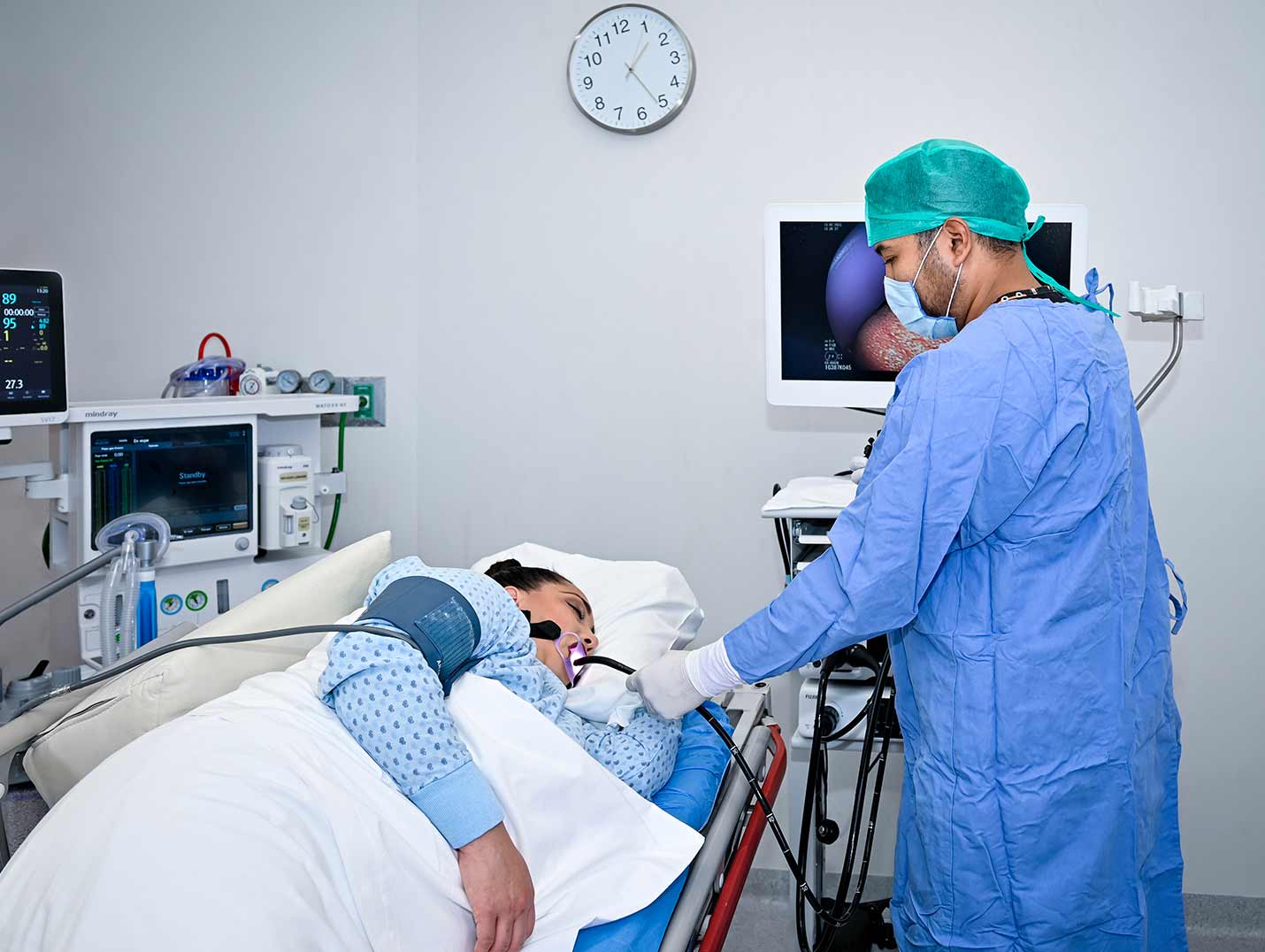 Doctor con pijama azul para cirugía, gorro verde y guantes blancos, realizando una endoscopia digestiva a una paciente