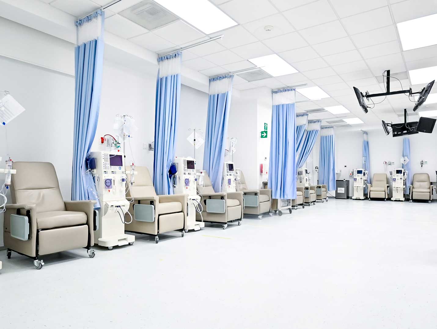 Sala de hemodiálisis con siete máquinas blancas, nueva sillones grises, cortinas azules y cinco pantallas colgadas del techo