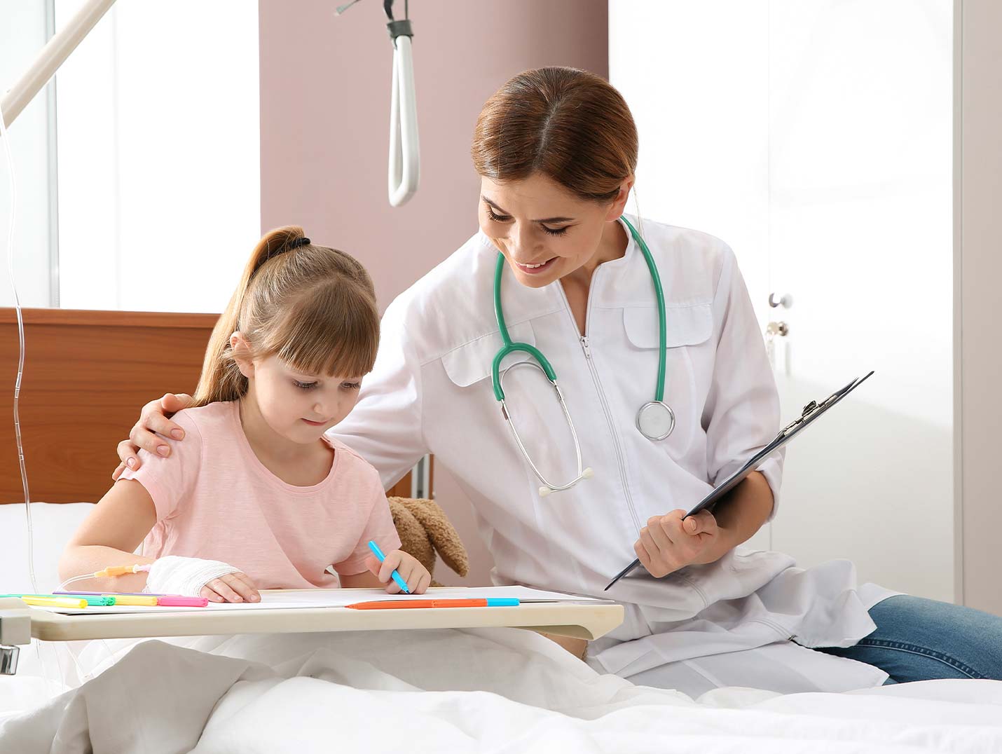 Niña sentada en una cama hospitalaria coloreando, mientras una doctora con bata blanca está a su lado tocando su hombro
