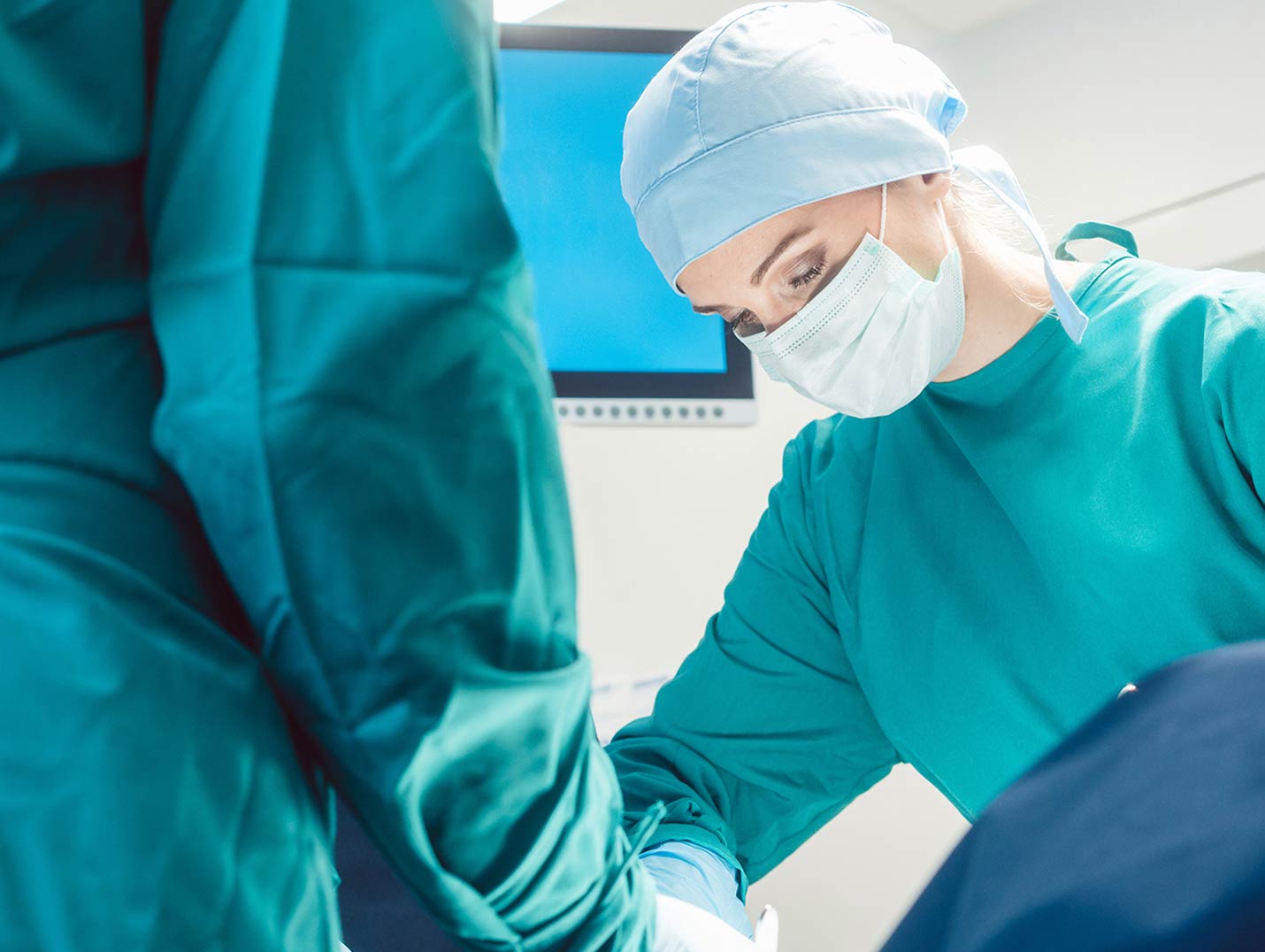Dos médicos con pijamas verdes para cirugía, gorro, cubrebocas y guantes azules, realizando un procedimiento quirúrgico