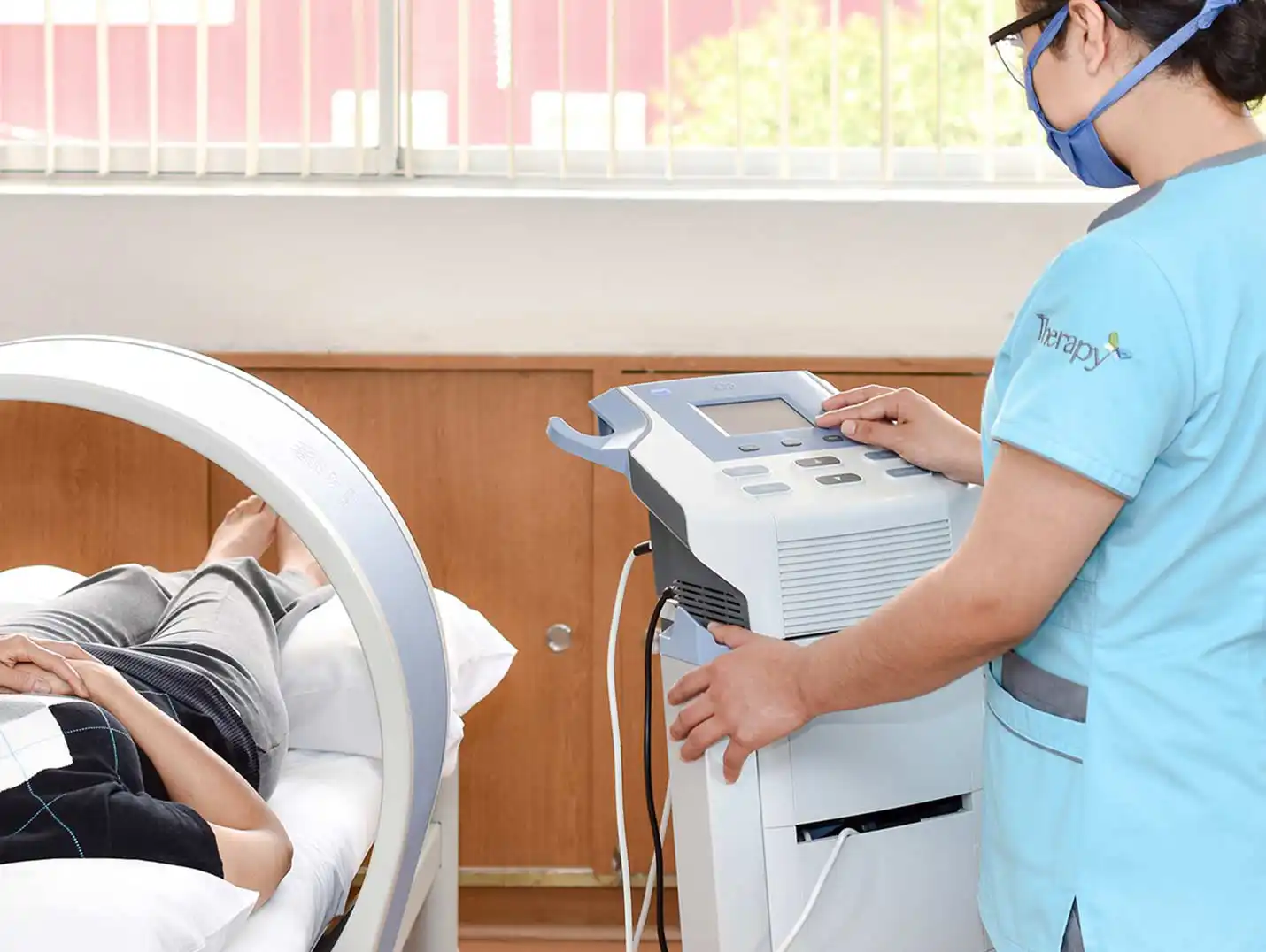 Paciente acostada en el equipo de magnetoterapia, mientras una terapeuta con uniforme médico azul lo controla