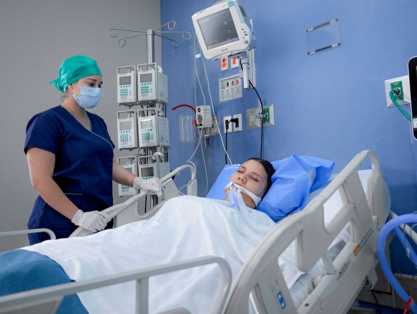 Paciente intubada y acostada en una cama del área terapia intensiva. A su lado se encuentra una doctora monitoreándola