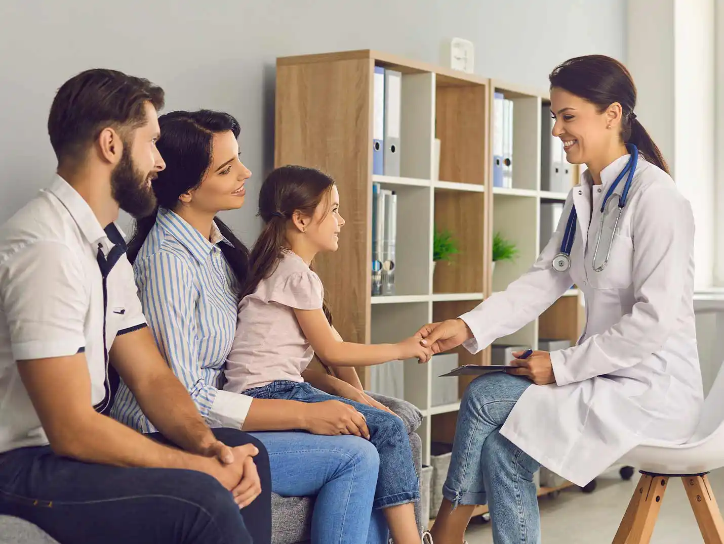 Padres e hija pequeña sentados en el sillón de un consultorio, mientras hablan con una doctora que usa bata blanca