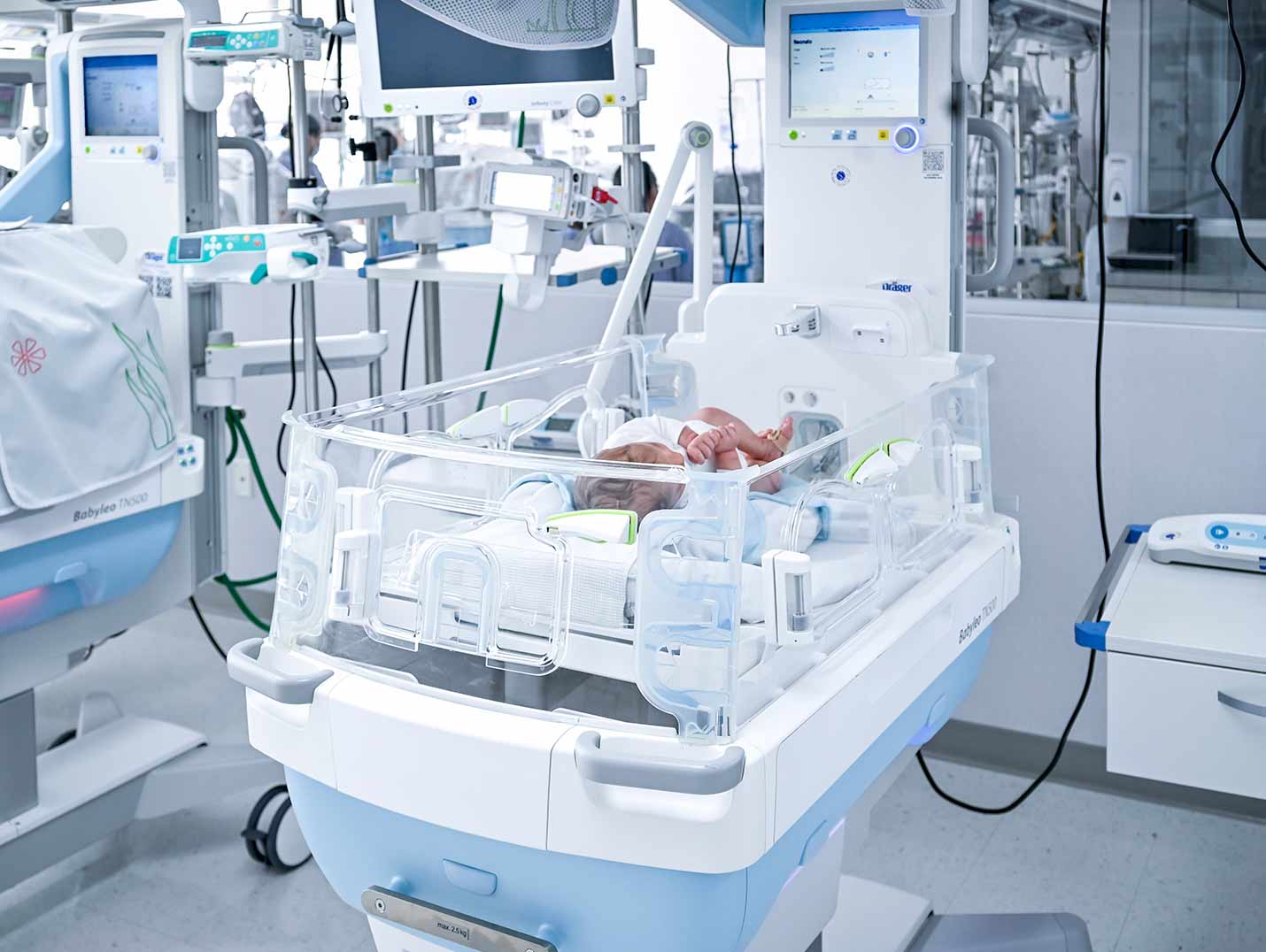 Bebé recién nacido dentro de una incubadora de color blanco y azul. A su lado hay un monitor de signos vitales