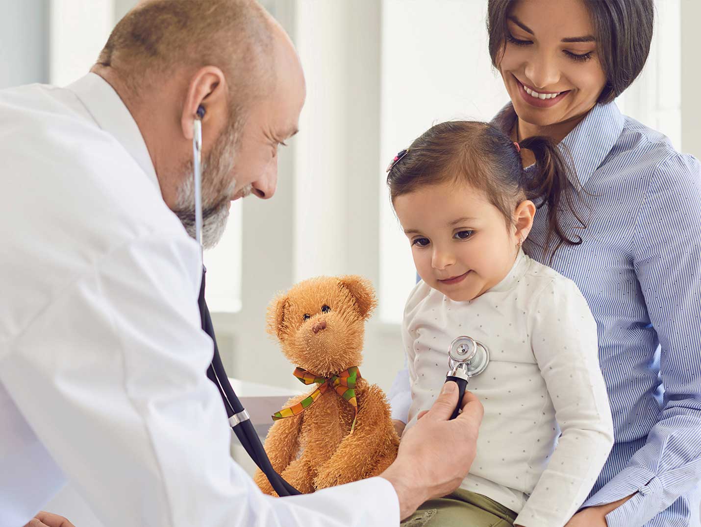 Doctor con bata blanca escuchando los latidos del corazón de una niña pequeña. A lado de ella está su mamá y un oso de peluche