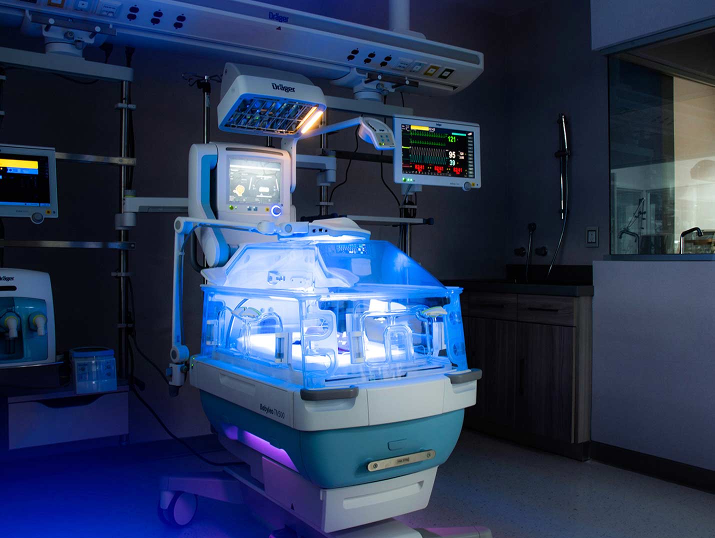 Área de terapia intensiva neonatal con una incubadora de color blanco y azul, alumbrada con luz de color azul