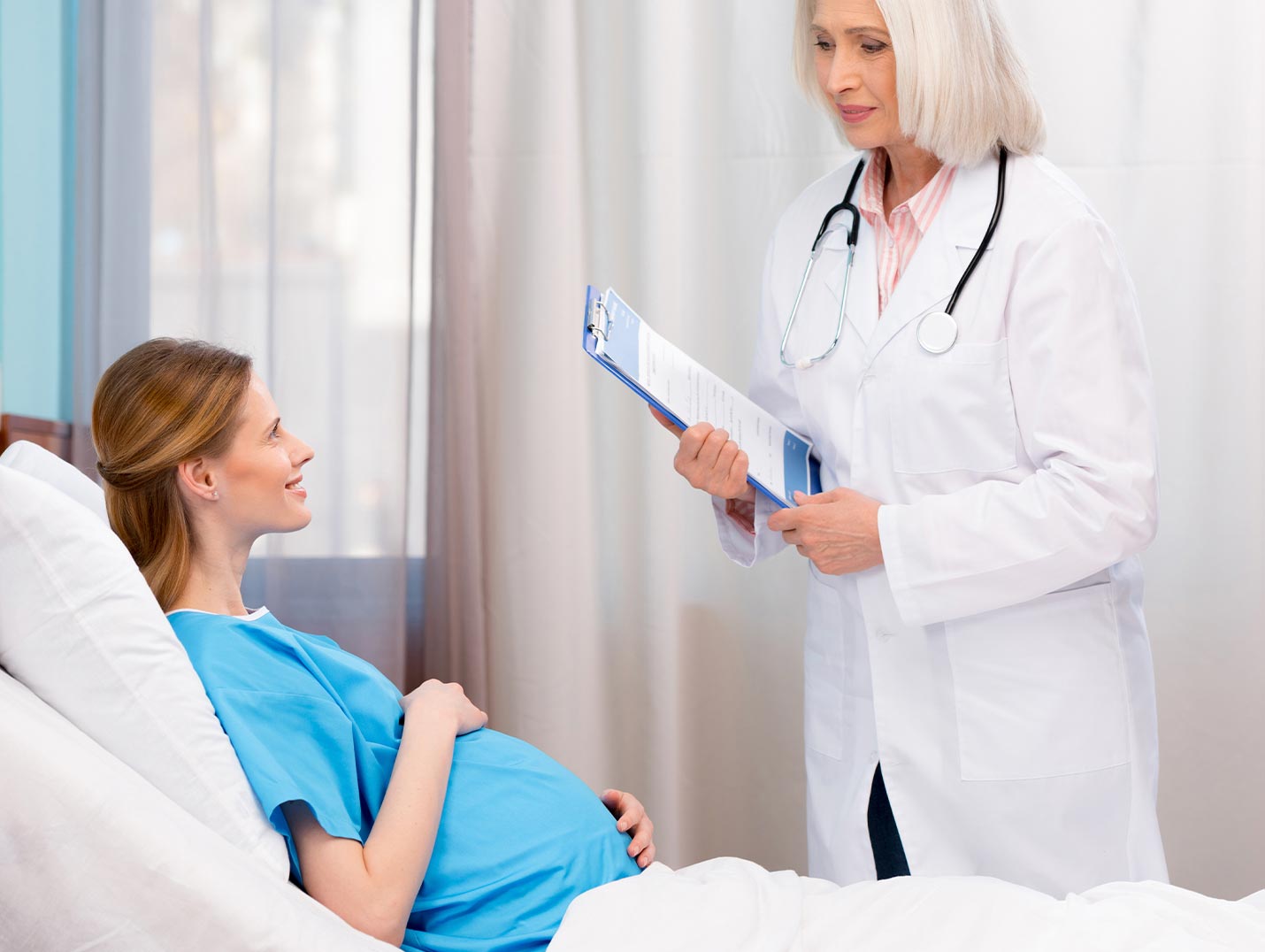 Doctora con bata blanca dando indicaciones a una paciente embaraza que está recostada en una cama hospitalaria