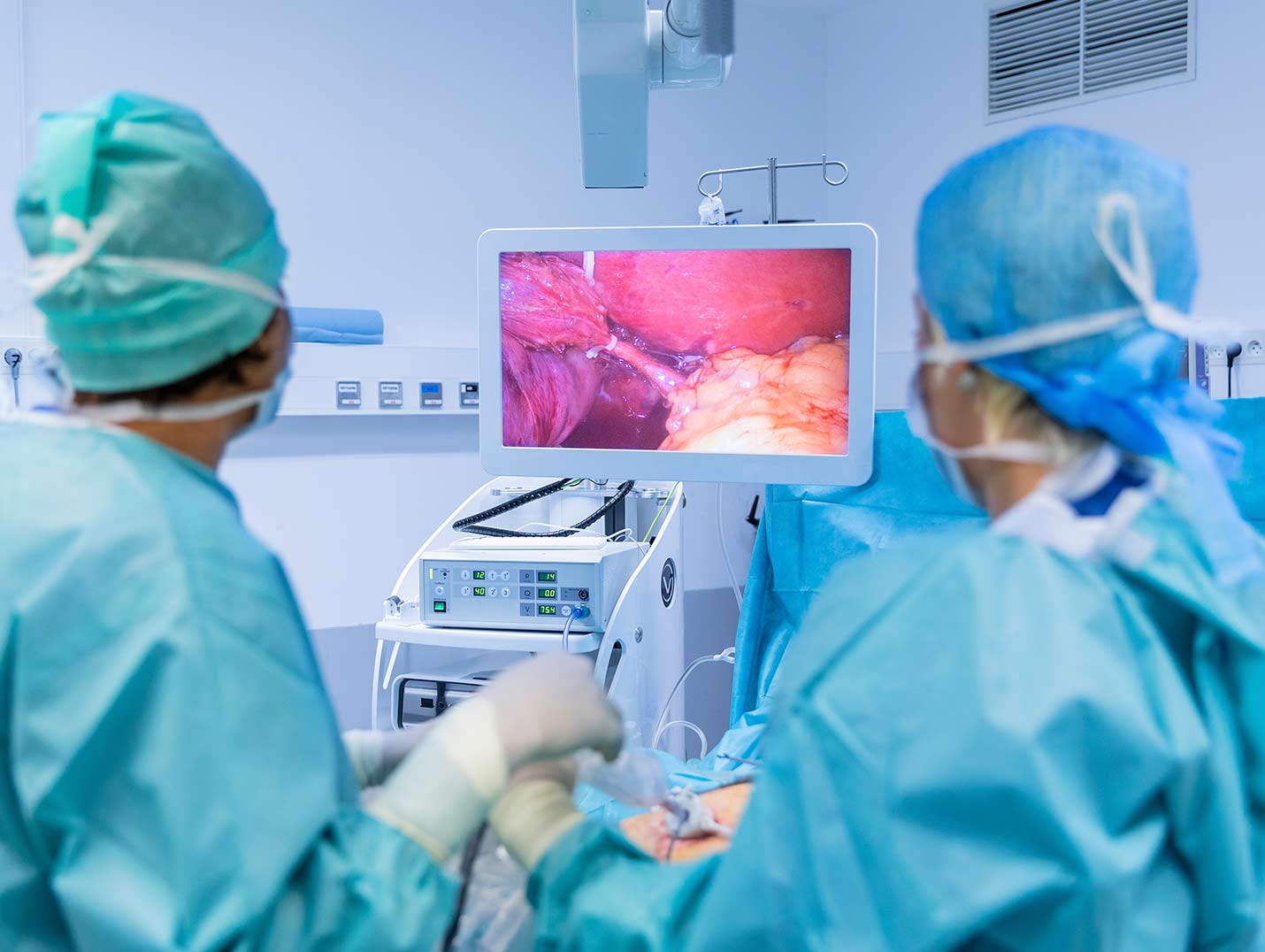 Dos médicos con pijamas azules para cirugía, gorros y guantes blancos, realizando una endoscopia y observando el monitor