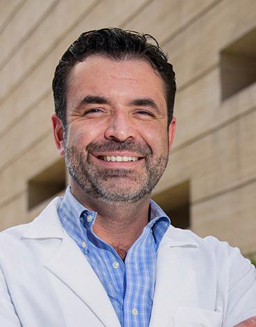 Es la foto de perfil de un ortopedista y traumatólogo en Ciudad de México de Hospital Angeles. El doctor viste una bata blanca y una camisa azul cielo de cuadros blancos, está sonriendo a la cámara. Es de tez caucásica, cabello negro y cejas pobladas.