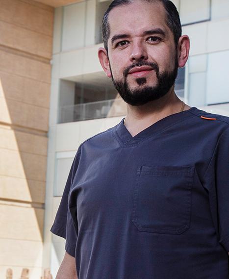 Es la foto de perfil de un otorrinolaringólogo en Ciudad de México de Hospital Angeles Clínica Londres. El doctor tiene el cabello oscuro y barba recortada. Está vestido con un uniforme médico de color gris oscuro y está de pie frente a un edificio moderno del hospital con amplias ventanas y una fachada de piedra clara.