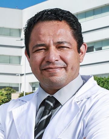 Es la foto de perfil de un internista y hematólogo en Ciudad de México de Hospital Angeles. Viste una bata blanca con una camisa gris y una corbata negra con rayas blancas, es de tez morena y está sonriendo suavemente a la cámara. 