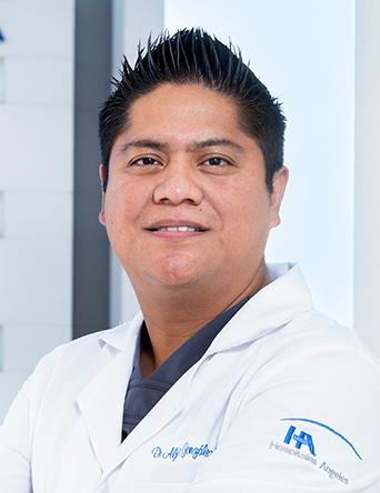 Es la foto de perfil de un ginecólogo y obstetra en Ciudad de México de Hospital Angeles Clínica Londres. El doctor tiene el cabello oscuro y corto, peinado hacia arriba. Está sonriendo y viste una bata médica blanca con su nombre y la especialidad bordados en el pecho. En la manga de la bata se distingue el logotipo del Hospital Ángeles. El fondo muestra una parte del edificio moderno del hospital.