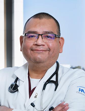 Es la foto de perfil de un nefrólogo en Ciudad de México de Hospital Angeles Clínica Londres. El doctor tiene el cabello corto y oscuro, y usa gafas. Está sonriendo y lleva puesto un estetoscopio alrededor del cuello. Viste una bata médica blanca con el logotipo del Hospital Ángeles en la manga. El fondo muestra una parte del edificio moderno del hospital y un cielo despejado.