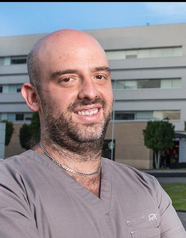 Es la foto de perfil de un ortopedista y traumatólogo en Ciudad de México de Hospital Angeles. El especialista viste un uniforme de color gris y una cadena plateada. Es de tez blanca, tiene barba y es calvo.