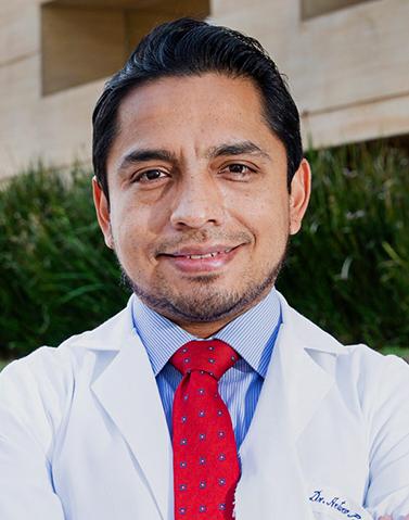 Es la foto de perfil de un cirujano general especialista en cirugía oncológica en Ciudad de México de Hospital Angeles. El doctor está sonriendo, viste una bata blanca, corbata roja con lunares azules con el centro blanco y una camisa rayada azul con blanco. 