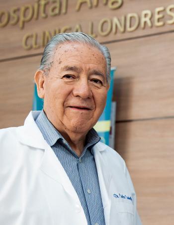 Es la foto de perfil de un cirujano neurológico en Ciudad de México de Hospital Angeles Clínica Londres. El doctor tiene el cabello canoso y lleva una expresión amigable. Viste una bata médica blanca sobre una camisa a rayas azules. El fondo muestra una pared de madera clara con el nombre del hospital en letras doradas.