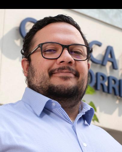 Es la foto de perfil de un ortopedista y traumatólogo en Ciudad de México de Hospital Angeles Clínica Londres. El doctor tiene el cabello oscuro y rizado, y usa gafas de montura negra. Lleva barba y bigote recortados. Viste una camisa de color azul claro y tiene una expresión relajada. El fondo muestra un edificio del hospital con un letrero en letras grandes.