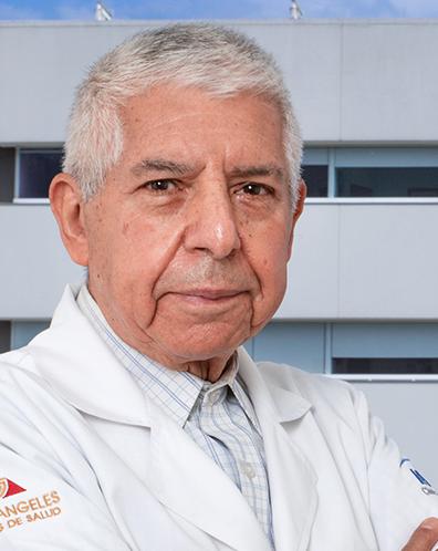 Es la foto de perfil de un cirujano general en Ciudad de México de Hospital Angeles Clínica Londres. El doctor tiene el cabello canoso y corto, y lleva una expresión seria. Viste una bata médica blanca sobre una camisa a cuadros claros. En la manga de la bata se distingue el logotipo de 'Grupo Ángeles Servicios de Salud'. El fondo muestra un edificio moderno del hospital con un cielo despejado.