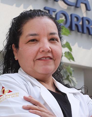 Es la foto de perfil de una geriatra en Ciudad de México de Hospital Angeles Clínica Londres. La doctora tiene el cabello oscuro y rizado, peinado hacia atrás. Viste una bata médica blanca sobre un uniforme negro. En la manga de la bata se distingue el logotipo de 'Grupo Ángeles Servicios de Salud'. Tiene una expresión amigable y el fondo muestra un edificio del hospital con un letrero en letras grandes.