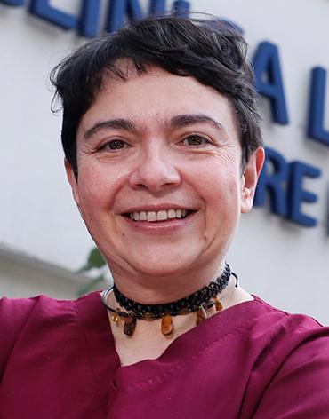 Es la foto de perfil de una ginecóloga y obstetra en Ciudad de México de Hospital Angeles Clínica Londres. La doctora tiene el cabello corto y oscuro, y está sonriendo. Viste un uniforme médico de color vino tinto y lleva un collar de cuentas negras con detalles marrones. El fondo muestra un edificio del hospital con un letrero en letras grandes.