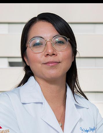 Es la foto de perfil de una hematóloga en Ciudad de México de Hospital Angeles Clínica Londres. La doctora tiene el cabello oscuro, lacio y suelto, y usa gafas de montura redonda. Viste una bata médica blanca con su nombre y la especialidad bordados en el pecho. En la manga de la bata se distingue el logotipo de 'Grupo Ángeles Servicios de Salud'. El fondo muestra una parte del edificio moderno del hospital.