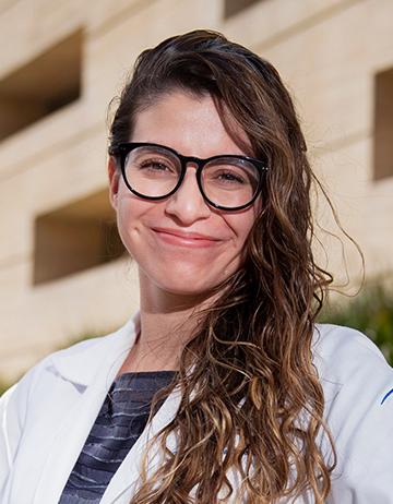 Es la foto de perfil de una dermatóloga en Ciudad de México de Hospital Angeles. La doctora tiene cabello ondulado, castaño claro, está sonriendo a la cámara. Tiene una bata blanca y una playera negra con rayas, usa lentes.