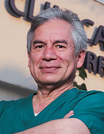 Es la foto de perfil de un cirujano neurológico en Ciudad de México de Hospital Angeles Clínica Londres. El doctor tiene el cabello canoso y corto, y está sonriendo ligeramente. Viste un uniforme médico de color verde. El fondo muestra un edificio del hospital con un letrero en letras grandes.