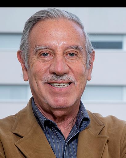 Es la foto de perfil de un cirujano neurológico en Ciudad de México de Hospital Angeles Clínica Londres. El doctor tiene el cabello canoso y un bigote. Está sonriendo y viste una chaqueta de color marrón sobre una camisa a rayas oscuras. El fondo muestra un edificio moderno del hospital con ventanas amplias.