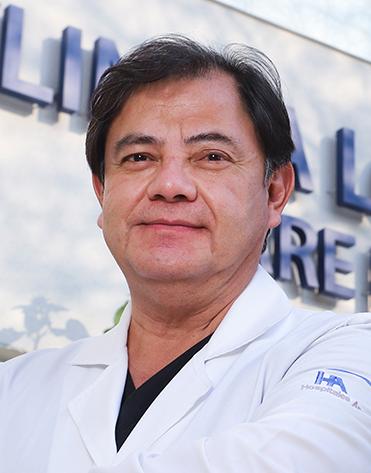 Es la foto de perfil de un neumólogo en Ciudad de México de Hospital Angeles Clínica Londres. El doctor tiene el cabello oscuro y corto, con una expresión calmada. Viste una bata médica blanca con el logotipo del Hospital Ángeles en la manga. El fondo muestra un edificio del hospital con un letrero en letras grandes.