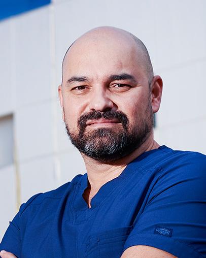 Es la foto de perfil de un cirujano oncológico en Ciudad de México de Hospital Angeles Lindavista. Es calvo. Tiene la barba y bigote negro. Usa un uniforme médico azul.