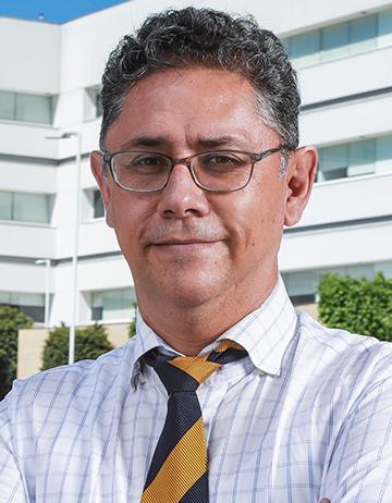 Es la foto de perfil de un cirujano general en Ciudad de México de Hospital Angeles. Viste una camisa de cuadros con una corbata azul marino con una franja ancha amarilla. El doctor tiene lentes y está sonriendo ligeramente.