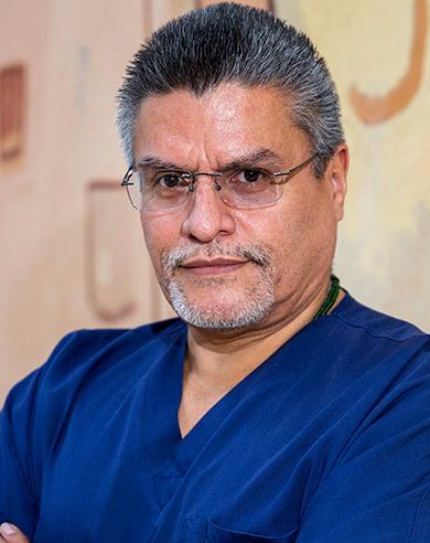 Es la foto de perfil de un cirujano plástico en Ciudad de México de Hospital Angeles Lindavista. Tiene el cabello y barba gris. Usa lentes y un uniforme médico azul. Tiene los brazos cruzados.