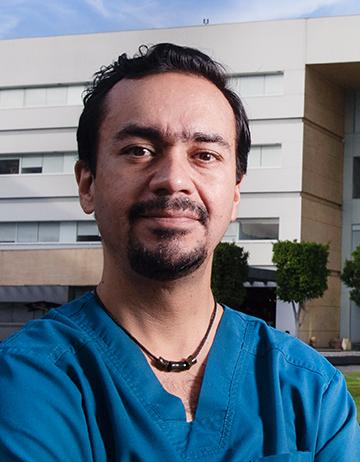 Es la foto de perfil de un ginecólogo y obstetra especialista en biología de reproducción en Ciudad de México de Hospital Angeles. Viste un uniforme azul, un collar de 3 anillos negro, tiene barba y el cabello ondulado. 