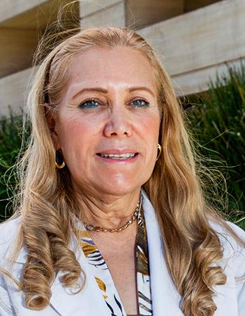 Es la foto de perfil de una dermatóloga en Ciudad de México de Hospital Angeles. La doctora viste una bata blanca, blusa de flores y joyería dorada, tiene el cabello rubio y en caireles.