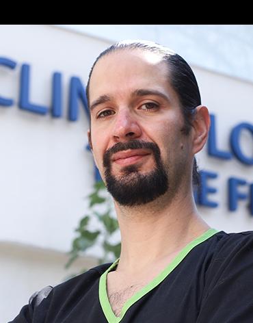 Es la foto de perfil de un cirujano plástico en Ciudad de México de Hospital Angeles Clínica Londres. Viste un uniforme oscuro con el cuello verde claro, tiene barba de candado, tez blanca y de fondo está el hospital.