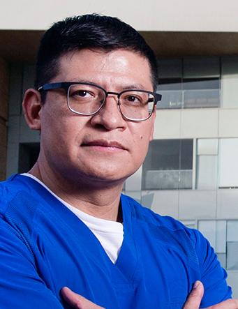 Es la foto de perfil de un cirujano oncológico en Ciudad de México de Hospital Angeles. Usa un uniforme azul con playera blanca y lentes con marco negro.