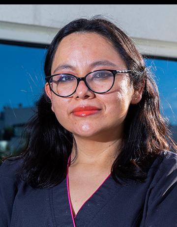 Es la foto de perfil de una endocrinóloga en Ciudad de México de Hospital Angeles Clínica Londres. La doctora tiene el cabello oscuro y lacio, usa gafas de montura negra y lleva labial rojo. Está sonriendo ligeramente y viste un uniforme médico de color negro con detalles en rosa. El fondo muestra un edificio con una ventana grande reflejando el cielo azul.