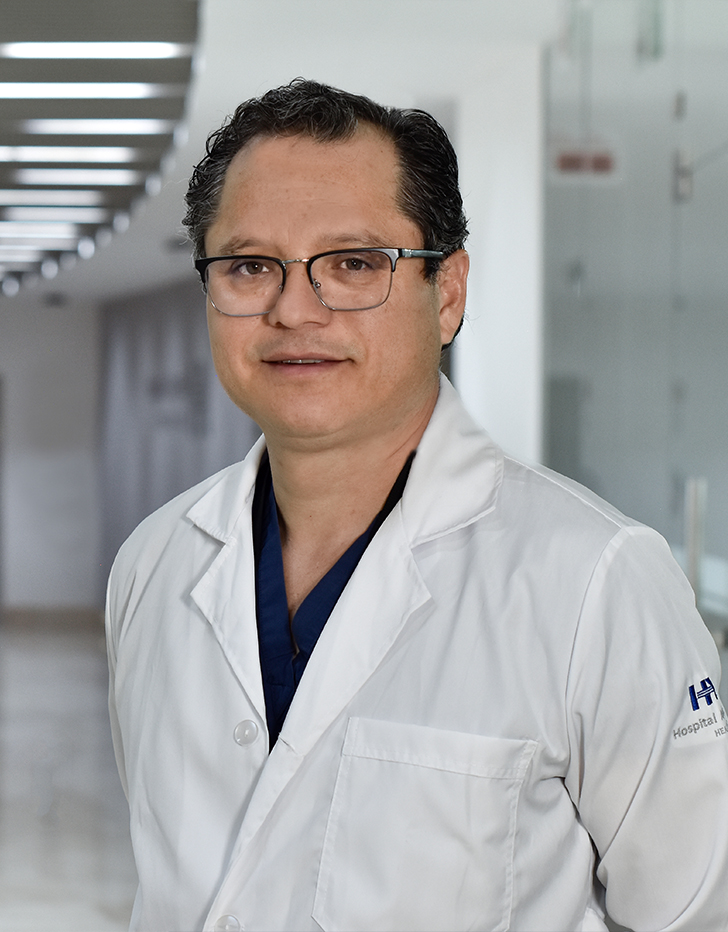 Es la foto de perfil de un cirujano cardiotorácico en Huixquilucan. Tiene el cabelo negro. Usa lentes, una bata blanca y un uniforme médico color azul.