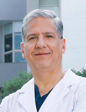 Es la foto de perfil de un coloproctólogo y cirujano genral en San Pedro Garza de Hospital Angeles Valle Oriente. Usa una bata blanca. Tiene el cabello gris y está sonriendo.