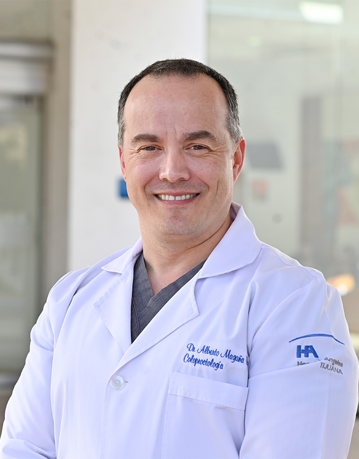 Es la foto de perfil de un cirujano general y coloproctólogo en Tijuana de Hospital Angeles Tijuana. Usa una bata blanca y un uniforme médico gris. Tiene el cabello negro y gris. Está sonriendo.