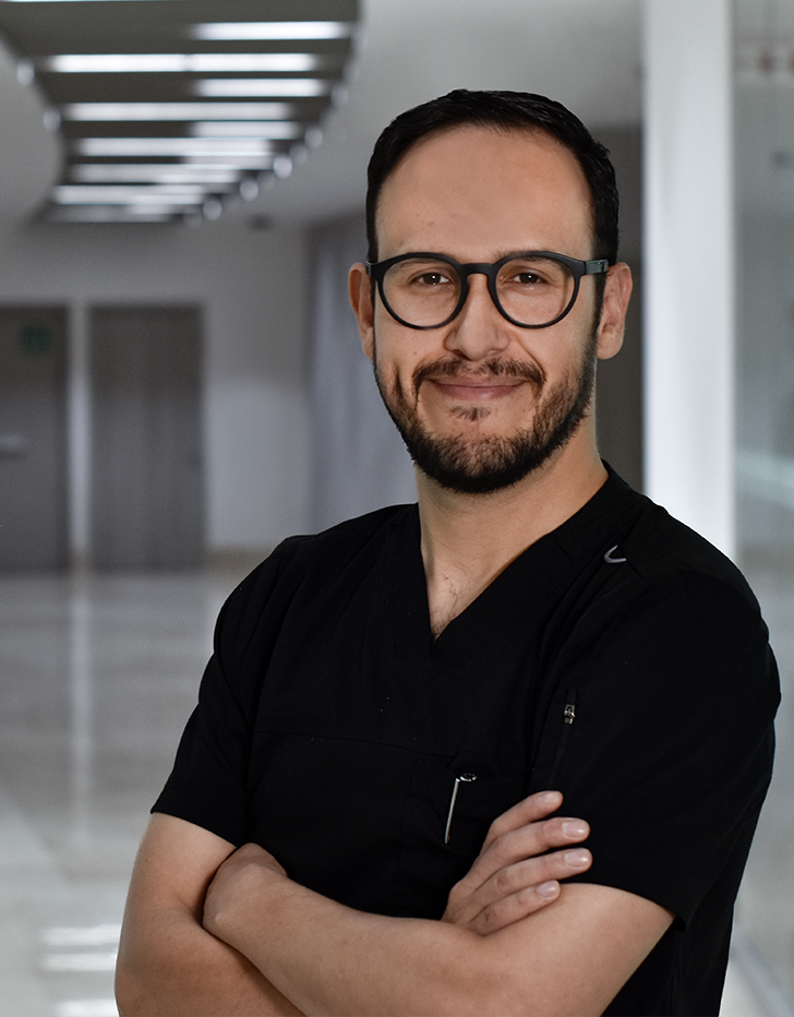 Es la foto de perfil de un cirujano pediátrico en Huixquilucan. Tiene el cabello negro. Usa lentes y un uniforme médico negro. Tiene los brazos cruzados y está sonriendo.