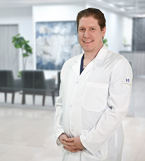 Es la foto de perfil de un terapeuta endovascular en neurología y cirujano neurológico en Ciudad de México de Hospital Angeles. Tiene un uniforme de color gris y de fondo se encuentra el hospital. 