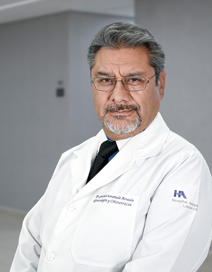 Es la foto de perfil de un ginecólogo y obstetra en Ciudad de México de Hospital Angeles Lindavista. Tiene el cabello y barba gris. Usa una bata blanca con una camisa azul. 
