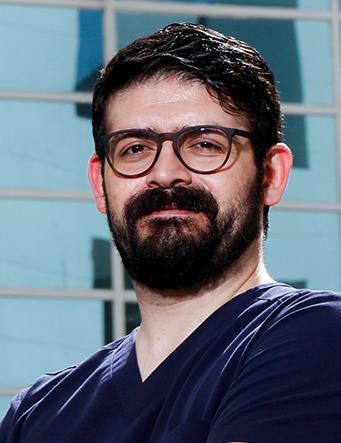 Es la foto de perfil de un cirujano general en San Pedro Garza de Hospital Angeles Valle Oriente. Usa un uniforme médico azul marino. Tiene el cabello y barba negra. Usa lentes.