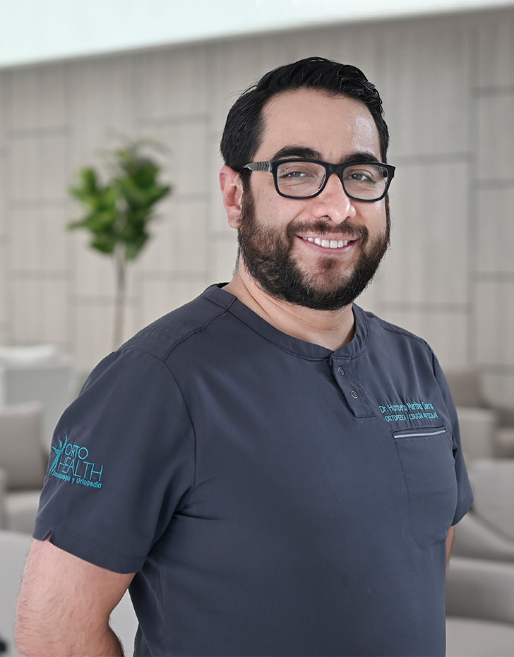 Es la foto de perfil de un cirujano general en Querétaro de Hospital Angeles Centro Sur. está sonriendo, viste un uniforme gris con letras bordadas azules, tiene barba y lentes con marco negro.