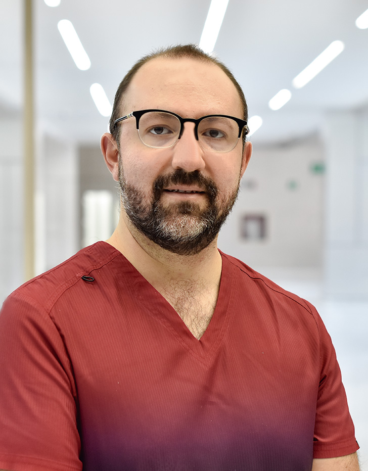 Es la foto de perfil de un cirujano pediátrico en Querétaro de Hospital Angeles Centro Sur. Viste un uniforme de color ladrillo, tiene barba y lentes.