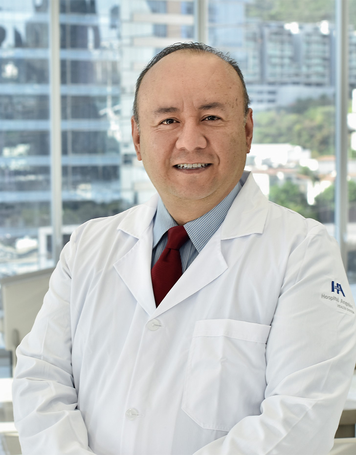 Es la foto de perfil de un cirujano plástico en San Pedro Garza de Hospital Angeles Valle Oriente. Usa una bata blanca con una camisa azul claro y una corbata vino. Está sonriendo y está sentado.